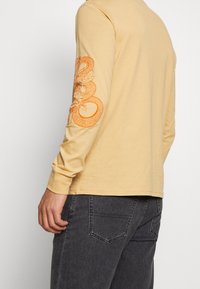 Langarmiges gelbes Hemd mit orangefarbenem Schlangendesign auf dem linken Ärmels, aus weichem Material, kombiniert mit dunkelgrauen Jeans.