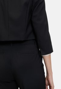 Giacca su misura nera con un design corto, maniche a tre quarti e una texture liscia. Abbinata a pantaloni su misura neri con tasche posteriori.