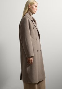Vrouw met lang blond haar, gekleed in een beige dubbelzijdig wollen jas en een bijpassende coltrui, staande zijwaarts tegen een eenvoudige muur.