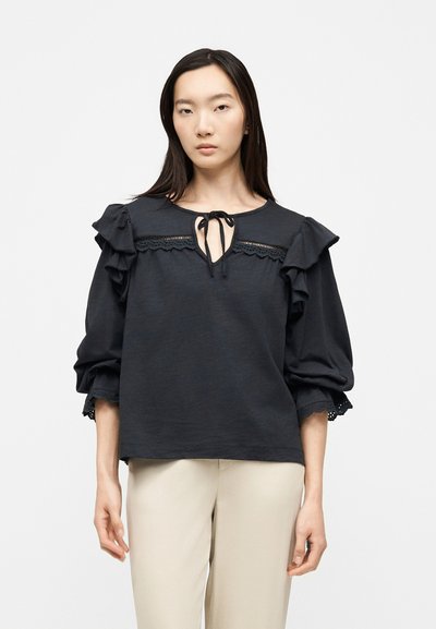 Femme portant un chemisier noir à épaules volantées, bordure en dentelle et col noué, associée à un pantalon beige clair, debout sur fond blanc.