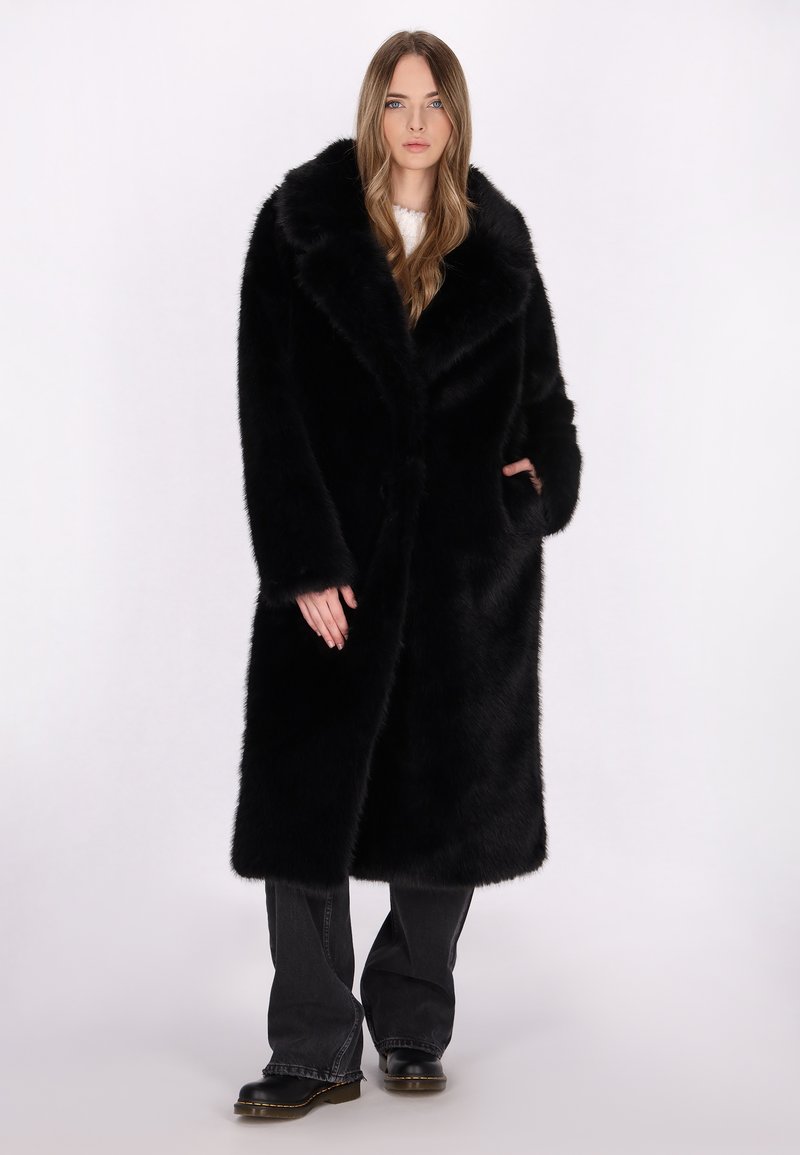 Lang sort faux fur frakke med en bred krave, enkelt knaplukning, to frontlommer og en glat tekstur. Båret over mørke bukser.