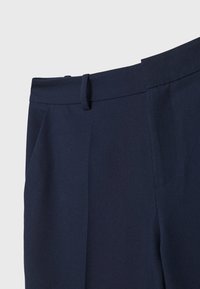 Gros plan d'un pantalon bleu foncé sur mesure montrant la ceinture, la boucle de ceinture et la couture de la poche latérale sur un fond uni.