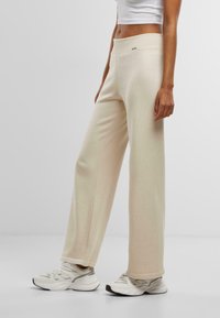 Pantaloni beige a gamba larga realizzati in un tessuto morbido con una texture liscia. Presentano un alto punto vita e una minima etichettatura sul davanti.