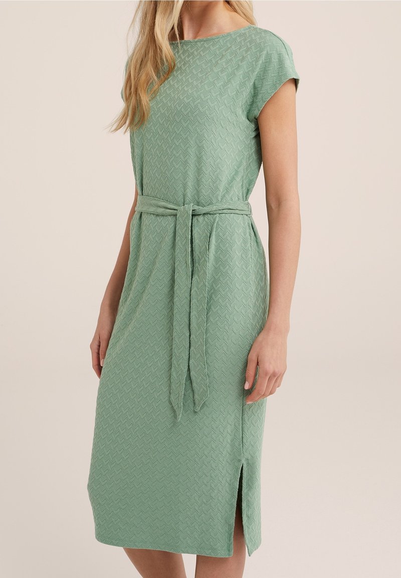 Vihreä, polvipituinen mekko, jossa on teksturoitu ziggzag-kuvio, lyhyet hihat ja yhteensopiva vyö vyötäröllä; sisältää sivuhiuksen mukavuutta varten.