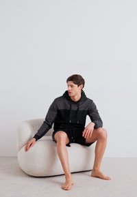 Black and gray hooded sweatshirt with patterned texture, worn with black shorts. Model seated on a curved, light-colored upholstered chair.