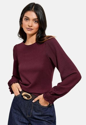 Hosszú, sötét hajú nő bordó színű, fodros mandzsettás hosszú ujjú felsőt, sötétkék farmert és fekete övet arany csattal visel.