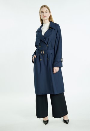 Marineblauwe trenchcoat met dubbele rij knopen, tailleceintuur en klepzakken. Gladde stof en brede mouwen, gecombineerd met zwarte broek met wijde pijpen.