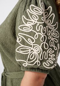 Olijfgroene jurk van textuurstof. Heeft witte borduursels op de mouw met spiraalvormige patronen. Inclusief een cinched taille.