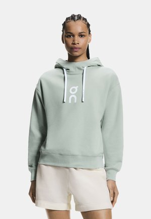 Kvinde med flettet hår iført en lysegrøn hættetrøje med hvide snore og beige shorts, stående mod en ensfarvet baggrund.