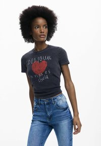 Noor naine loodusliku afro-soenguga, kellal must graafiline t-särk ja sinised tikanditega kõrge vöökohaga teksad, valgel taustal.