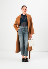 Femme portant un manteau beige sur un haut en dentelle bleu marine, un jean gris taille haute, des mocassins noirs, tenant un petit sac à main blanc sur un fond blanc.