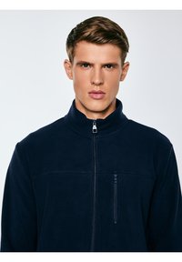 Młody mężczyzna o krótkich brązowych włosach, ubrany w granatową polarową kurtkę na zamek, na tle jasnego, jednolitego tła.