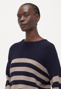 Mørkeblå strikkegenser med brede beige striper, rund hals og senkede skuldre. Teksturlignende stoff med løs passform.