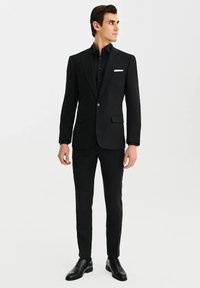 Czarny garnitur z satynowym klapą, dopasowana marynarka i spodnie proste. Biała chusteczka w kieszeni jako akcent, zestawiona z czarną koszulą i butami.