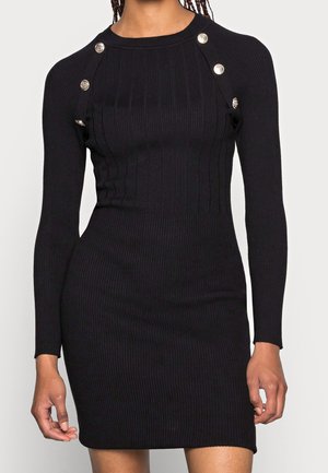 Femme portant une robe noire moulante à manches longues côtelée avec des boutons décoratifs argentés sur les épaules.