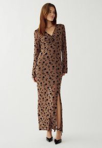 Dlhé hnedé šaty z elastického materiálu s čiernym kvetinovým vzorom, hlbokým výstrihom do V, dlhými rukávmi, rozparkom po stehná a priliehavým strihom.