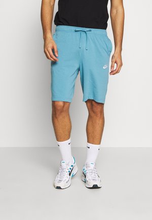Homme portant un short Nike bleu clair avec cordon, des chaussettes Nike blanches et des baskets blanches avec des accents bleus, debout sur un sol gris.