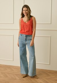 Woman standing smiling, wearing an orange lace-trimmed sleeveless top, light blue wide-leg jeans, and beige pointed shoes.