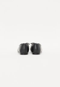 Ballerines en cuir noir avec un bout rond, accent de nœud noué à l'arrière et semelle en caoutchouc plate, présentées de dos sur un fond blanc.