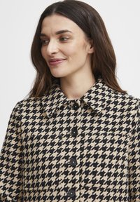 Bež ja mustriga houndstooth mantel, millel on krae, nööbiga sulgemine ja rinna tasku. Kanga pinnal on tekstuur.