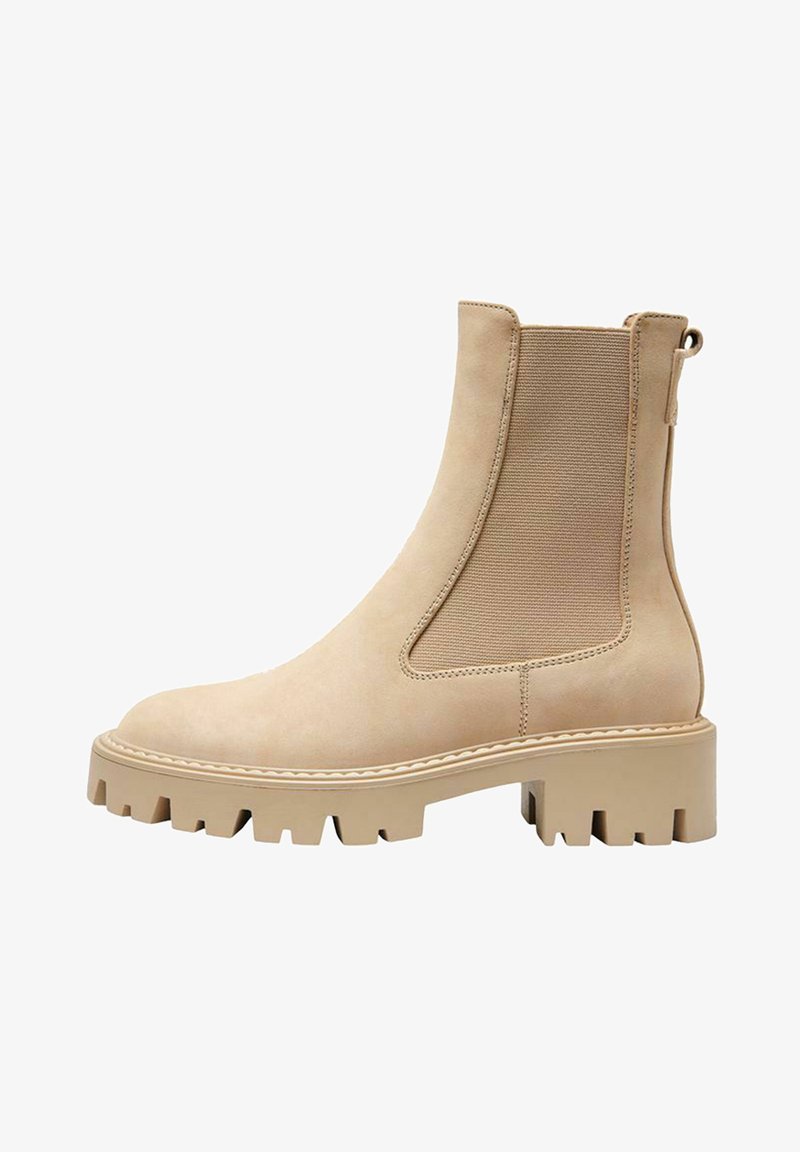 Bottines beiges avec une finition en suède lisse, des panneaux latéraux élastiques et une semelle crantée épaisse pour une traction optimale. Présente un détail de couture décoratif.