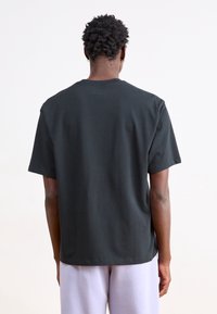 Osoba z krótkimi kręconymi włosami ubrana w gładką czarną koszulkę i jasne spodnie, stojąca przodem do białego tła.