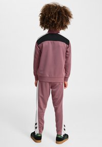 Dres dla dzieci w miękkim mauve z czarnym panelem na ramieniu, białymi paskami po bokach i czarnymi detalami w kształcie chevron na spodniach. Posiada wysoki kołnierz.