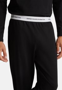 Zwarte joggingsbroek heeft een zachte, gladde textuur met een witte elastische tailleband waarop "UNITED COLORS OF BENETTON" is gedrukt.