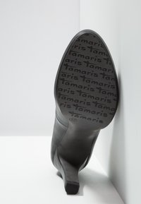 Chaussure à talons noirs inclinée pour montrer la semelle avec le logo "Tamaris" répété et la pointure 37, appuyée contre un coin de mur blanc.