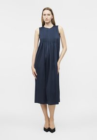 Γυναίκα που φοράει μπλε σκούρο midi φόρεμα χωρίς μανίκια με πλισέ μπούστο, και μαύρες μυτερές γόβες, στέκεται μπροστά σε λευκό φόντο.