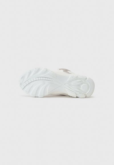 Semelle d'une sneaker en caoutchouc blanc, présentant un design texturé avec des rainures courbes et un logo en relief sur le côté.