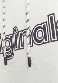 Sudadera gris claro con la impresión en negrita de "originals" en negro. Presenta una capucha con cordones a rayas en blanco y negro y un sutil logo debajo.