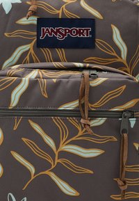 Sac à dos marron avec un motif de feuilles crème et doré ; il comporte une étiquette navy avec "JANSPORT", des tirettes de fermeture éclair marron et un tissu texturé.