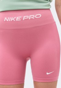 Shorts de sport roses moulants Nike Pro avec logo blanc et swoosh, portés par une personne avec un haut vert visible à la taille.