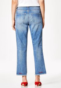 Světlé modré džíny se rovnýma nohama, roztrhaným lemem a kontrastním červeným vyšitým textem blízko lemu, s dvěma zadními kapsami.