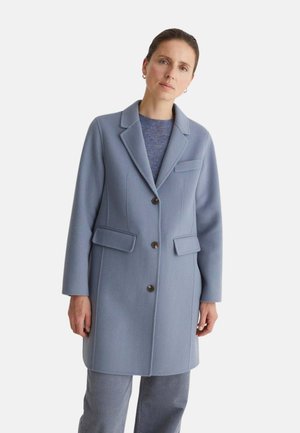 Mujer que lleva un abrigo largo azul claro con botones, bolsillos con solapa y cuello con muesca, combinado con unos pantalones azules a juego y un suéter.