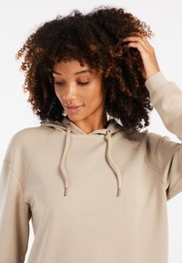 Felpa beige con cappuccio e lacci, con dettagli metallici, caratterizzata da una vestibilità rilassata, maniche raglan e una texture liscia e morbida.