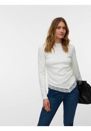 Femme aux cheveux châtain clair ondulés, portant un haut blanc à manches longues avec un ourlet en dentelle, jean bleu, tenant un sac à main noir sur fond blanc.