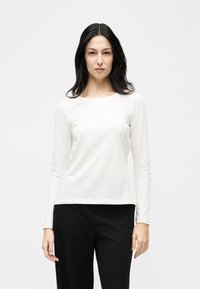 Γυναίκα με μακριά σκούρα μαλλιά που φοράει απλό λευκό μπλουζάκι με μακριά μανίκια και μαύρο παντελόνι, στέκεται μπροστά σε απλό λευκό φόντο.