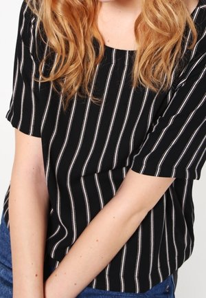 Žena s dlouhými vlnitými blond vlasy v černém tričku s krátkými rukávy a bílými svislými pruhy a modrými džínami, s rukama zkříženýma před sebou.