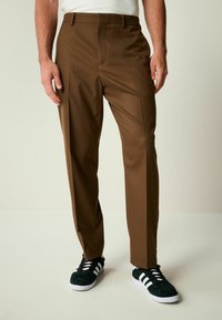 Pantaloni marroni su misura con pieghe frontali, una texture liscia e una vestibilità dritta, abbinati a sneaker nere con strisce bianche.