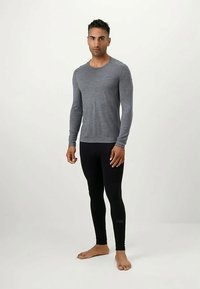 Άνδρας που στέκεται ξυπόλητος και φοράει ένα γκρι μακρυμάνικο πουκάμισο και μαύρα κολάν σε μια απλή φωτεινή φόντο.