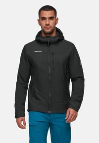 Uomo che indossa una giacca con cappuccio Mammut nera con tasche con zip e pantaloni cargo blu brillante, in piedi su uno sfondo bianco neutro.