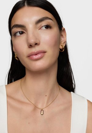 Frau mit dunklem Haar, die goldene Creolen und eine goldene Halskette mit einem schwarz umrandeten ovalen Anhänger trägt, gekleidet in ein weißes ärmelloses Top.