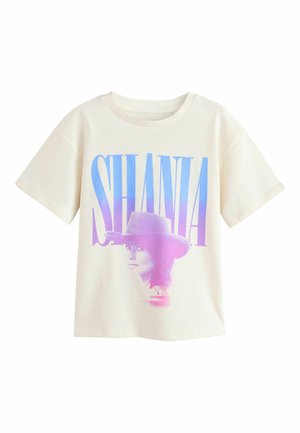 Junges Mädchen mit halbhochgestecktem Haar, trägt ein übergroßes weißes T-Shirt mit dem Schriftzug "SHANIA" und Grafik, weite Jeans und glitzernde beige Ballerinas.