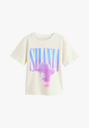 Junges Mädchen mit halbhochgestecktem Haar, trägt ein übergroßes weißes T-Shirt mit dem Schriftzug "SHANIA" und Grafik, weite Jeans und glitzernde beige Ballerinas.