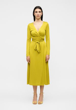 Γυναίκα που φοράει μακρυμάνικο μίντι φόρεμα τύπου μπολερό σε μουσταρδί χρώμα, δεμένο στη μέση, όρθια με γυμνά πόδια σε μονόχρωμο άσπρο φόντο.