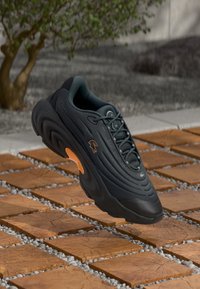 Sort og mørkegrå sneakers med orange sål detajle, svævende over en træblok belægning med grønt i baggrunden.