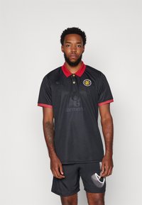 Zwarte poloshirt met een rode kraag en mouwen, voorzien van een logo op de linkerborst en een subtiele textuur. Gepaard met zwarte shorts.