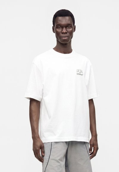 Άνδρας που φοράει λευκό υπερμεγέθες T-shirt με μικρό λογότυπο Off-White στο στήθος και ανοιχτό γκρι σορτς σε απλό φόντο.